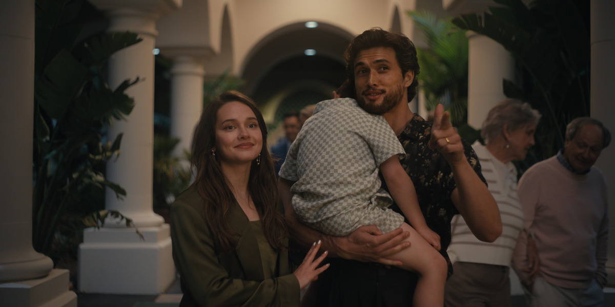 A smiling couple walks under archways surrounded by columns and greenery at night; the man is holding a child, and several other people are walking in the background, creating a warm, lively atmosphere.