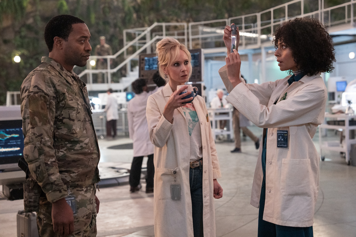 Chiwetel Ejiofor as Strickland wears camouflage while Juno Temple as Dr. Teddy Paine and Clark Backo as Sadie wear lab coats. All three stand in a circle observing a beaker in a still image from the film ‘Venom: The Last Dance.’