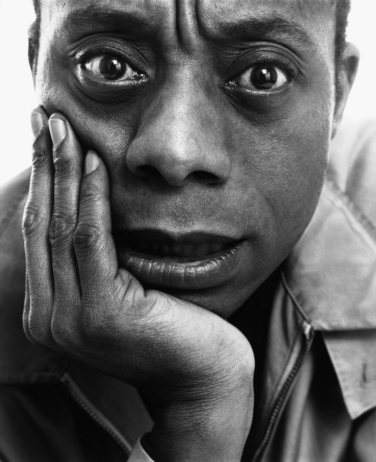 Black and white close-up portrait of a person resting their face on their hand, looking directly at the camera with an intense, contemplative expression, set against a plain background.