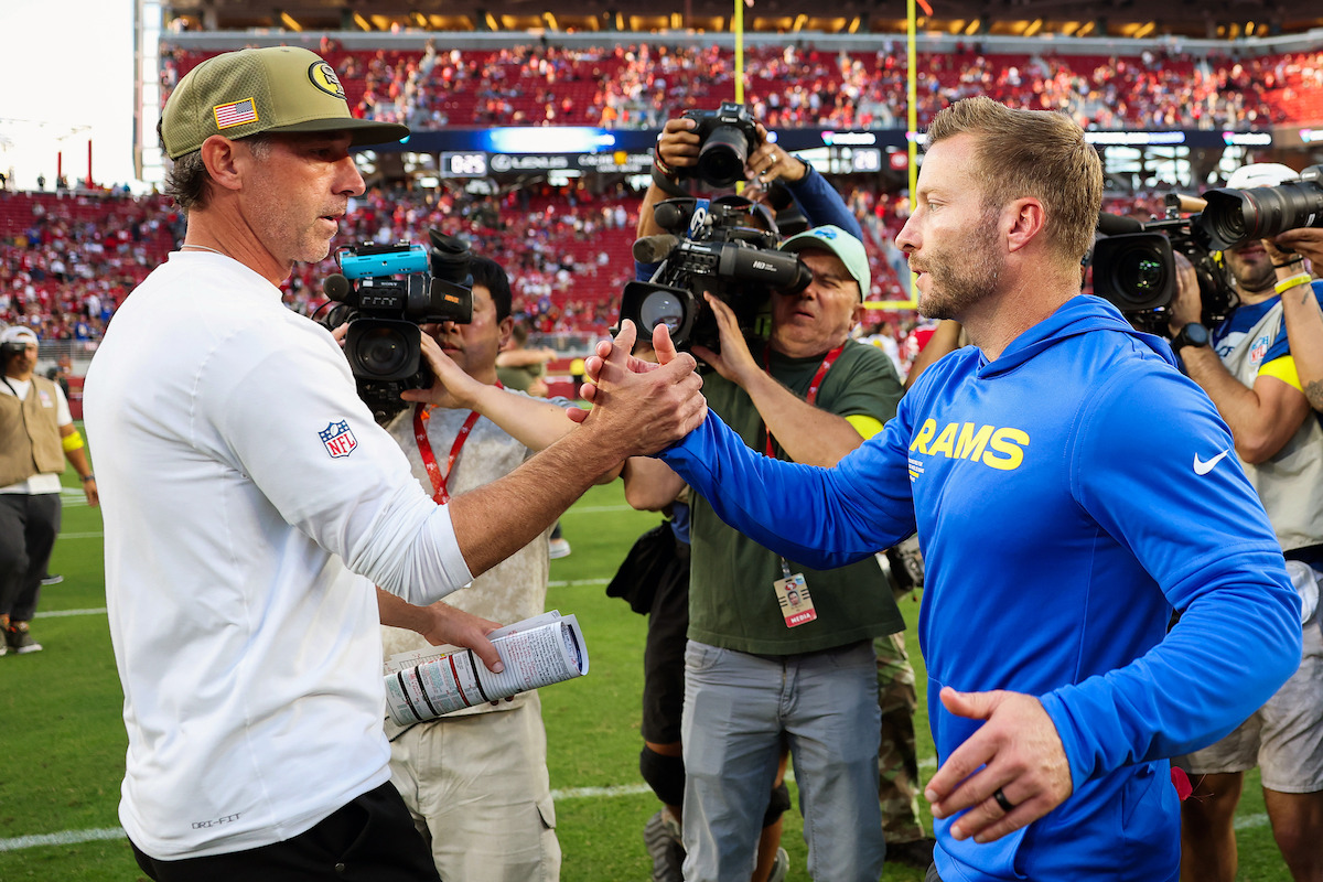 Two NFL coaches shake hands on a football field after a game, surrounded by photographers and cameras with a stadium full of fans in the background.