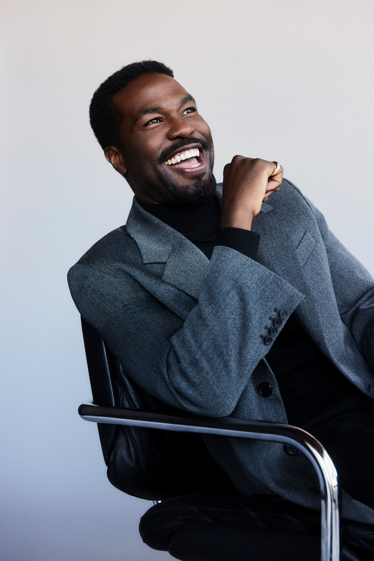 Smiling man in a gray blazer sitting on a black chair in a minimalist studio with a light background, looking relaxed and joyful.