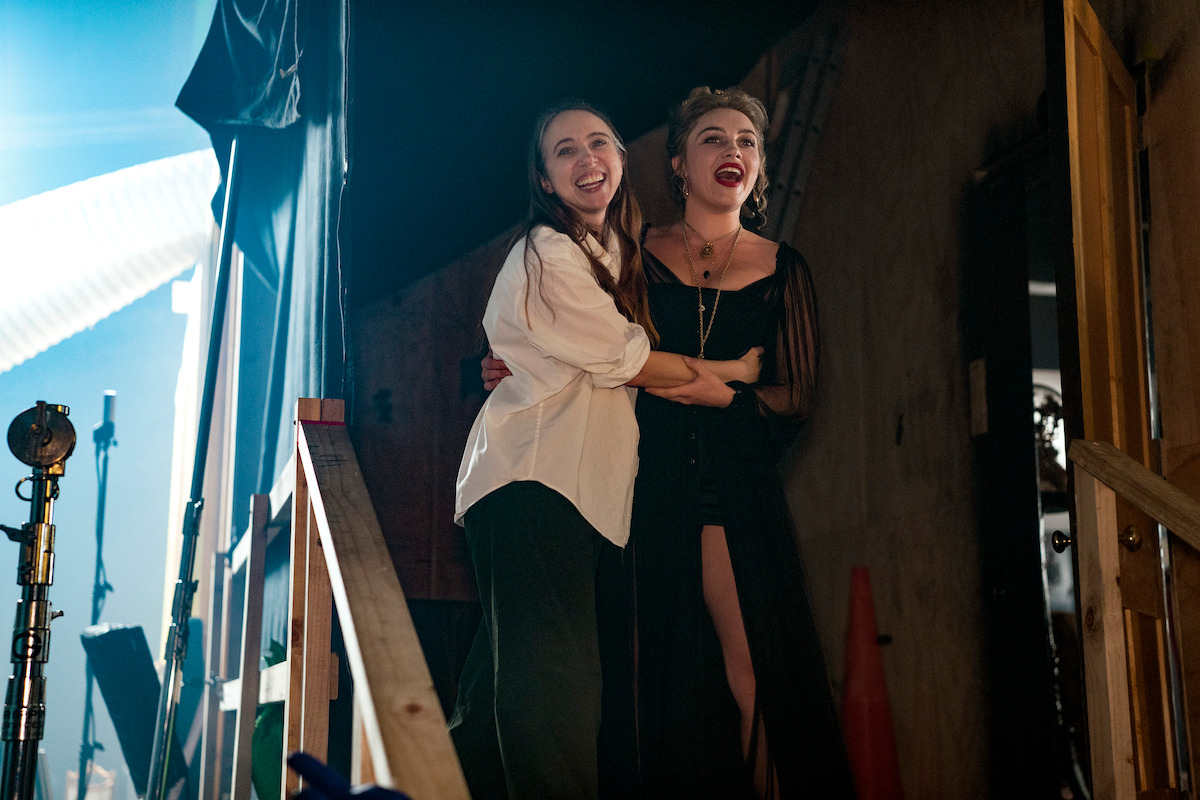 Two women stand backstage, embracing and smiling, surrounded by stage equipment, props, and dramatic lighting, conveying excitement and anticipation in a theater or performance setting.