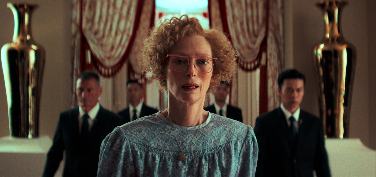 A woman in a blue dress stands before four men in suits in a lavish hotel room.