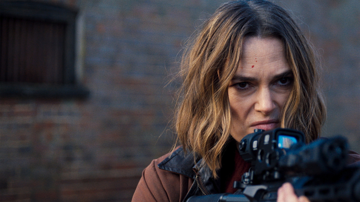 Mulher com cabelo na altura dos ombros, focada e atenta, apontando um rifle para a frente em um ambiente urbano perto de uma parede de tijolos.