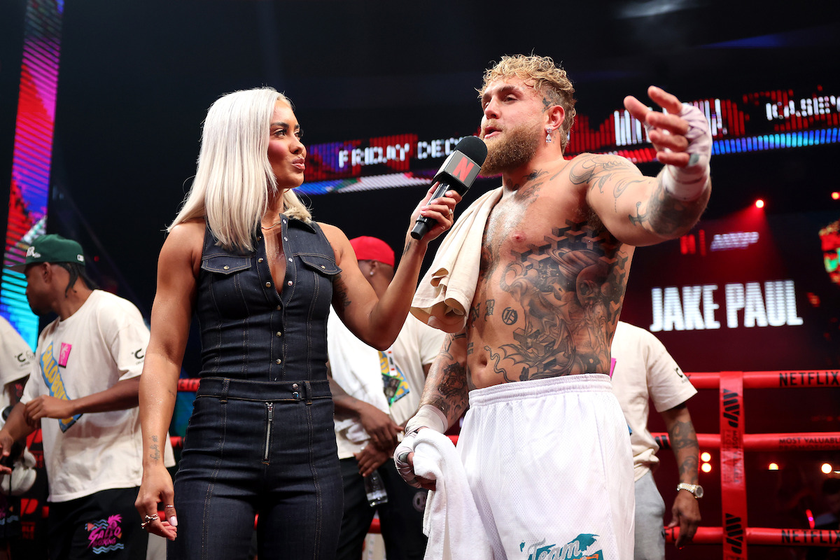 Jake Paul being interviewed by Sibley Scoles in a boxing ring, surrounded by people, with bright lights and signs in the background.