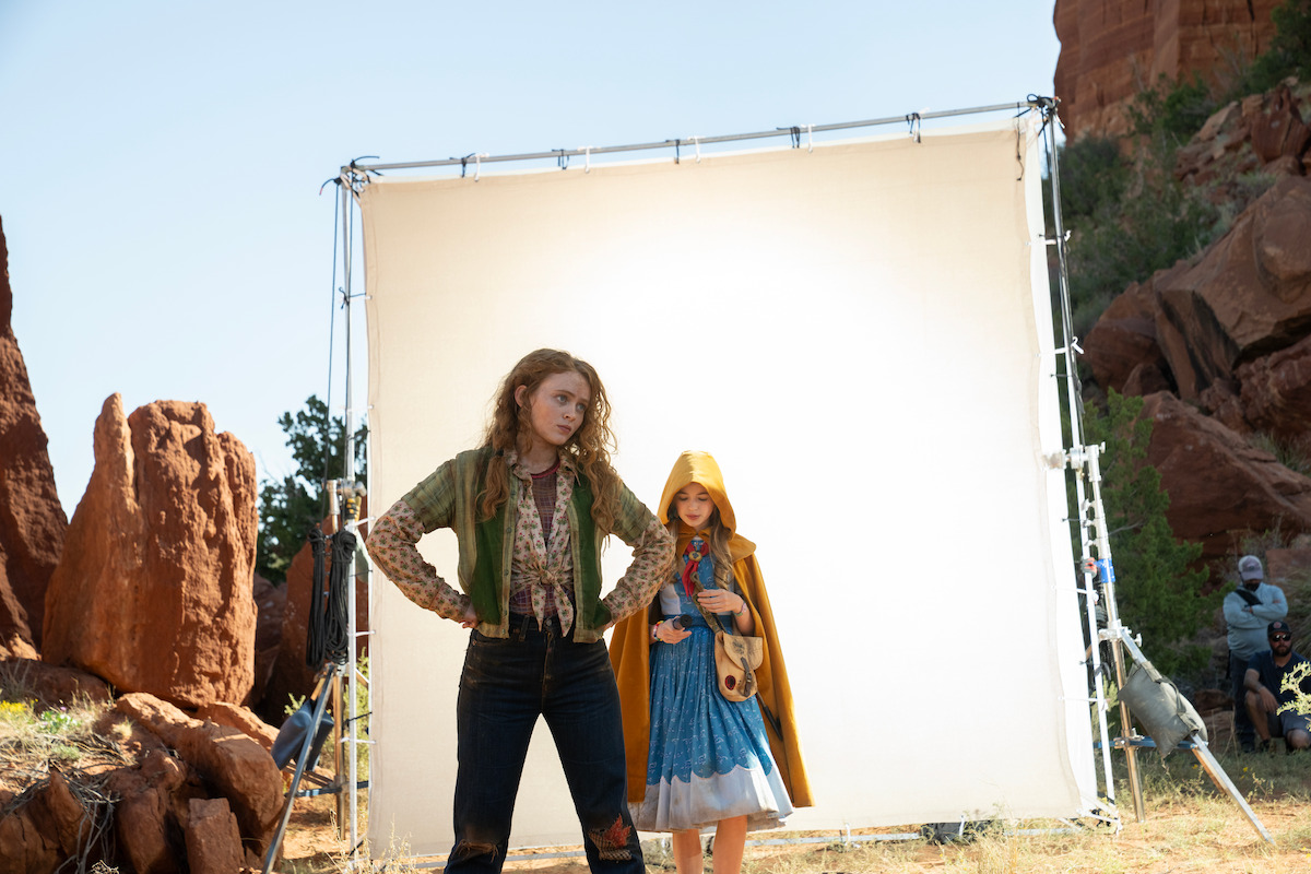Two girls in costume stand before a large white backdrop on a rocky outdoor film set.