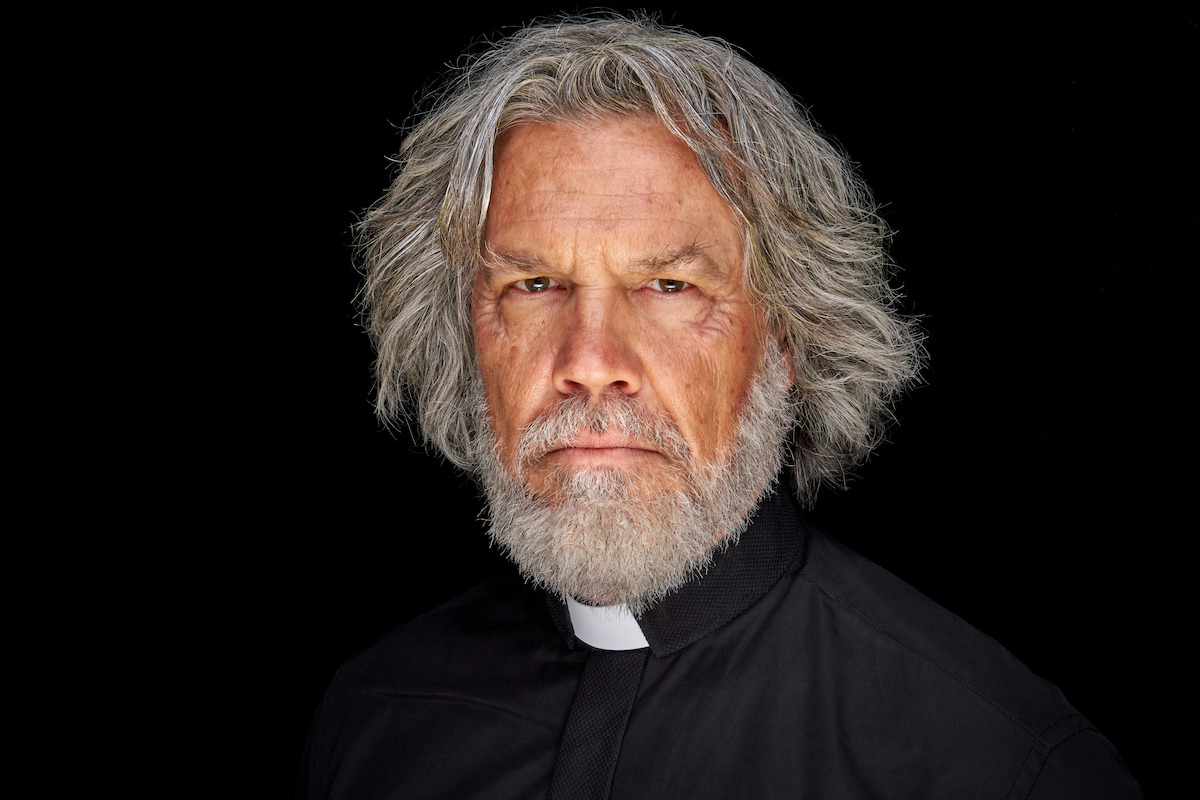 Older man with gray hair and beard, wearing a black clerical shirt and white collar, standing against a black background, looking serious and contemplative with a stern expression. Studio lighting, solemn mood.