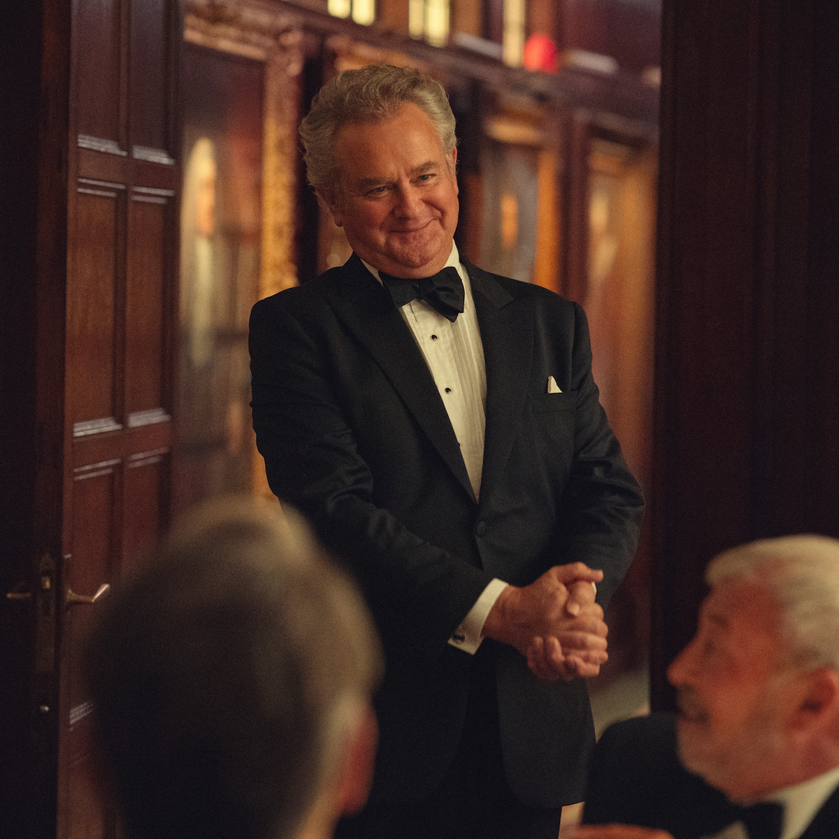 Man in a tuxedo smiling and standing in an elegant, wood-paneled room with portraits, while others in formalwear sit and interact in the foreground.