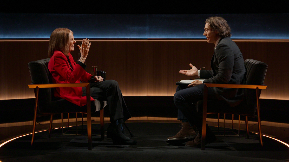 Two people seated facing each other in armchairs having a conversation in a dimly lit studio with wooden paneling and a dark background, creating a formal interview or talk show setting.