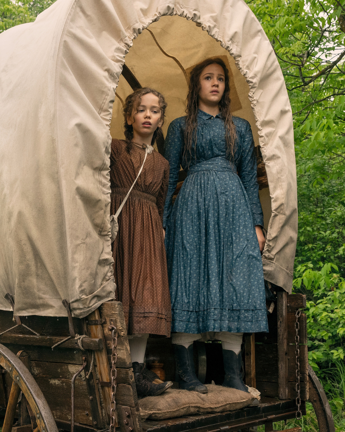 Two girls in pioneer era dresses stand inside a covered wooden wagon, surrounded by lush green foliage, looking outward with serious expressions, evoking a historical frontier setting.