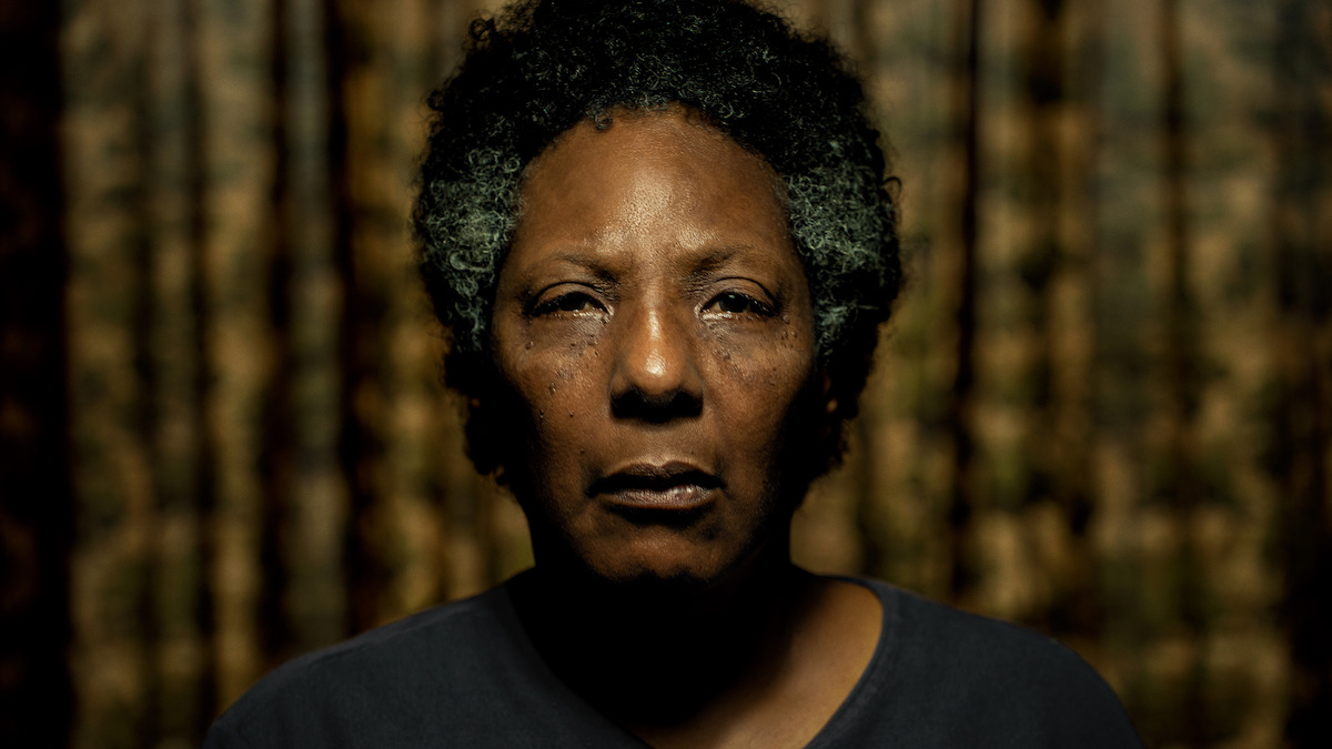 Imagem mostra uma mulher negra idosa em close, com os cabelos parcialmente brancos à mostra e a expressão séria