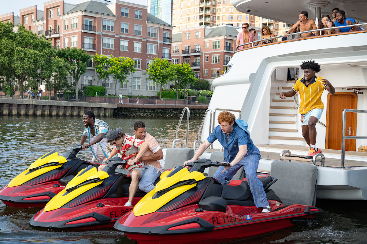 People prepare to ride jet skis off a yacht docked by the water, with a lively group watching from the deck and city buildings in the background, creating an energetic, urban summer scene.