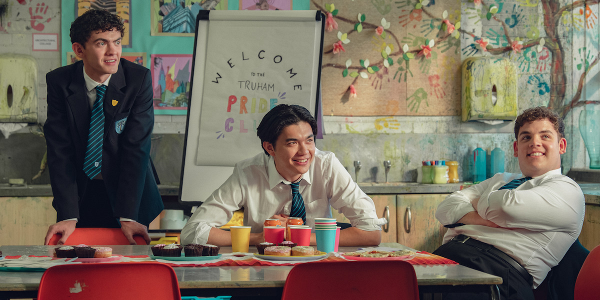 Three students in school uniforms sit and smile around a table with snacks and drinks in a colorful classroom decorated with children's artwork and a "Welcome to the Truham Pride Club" sign in the background.