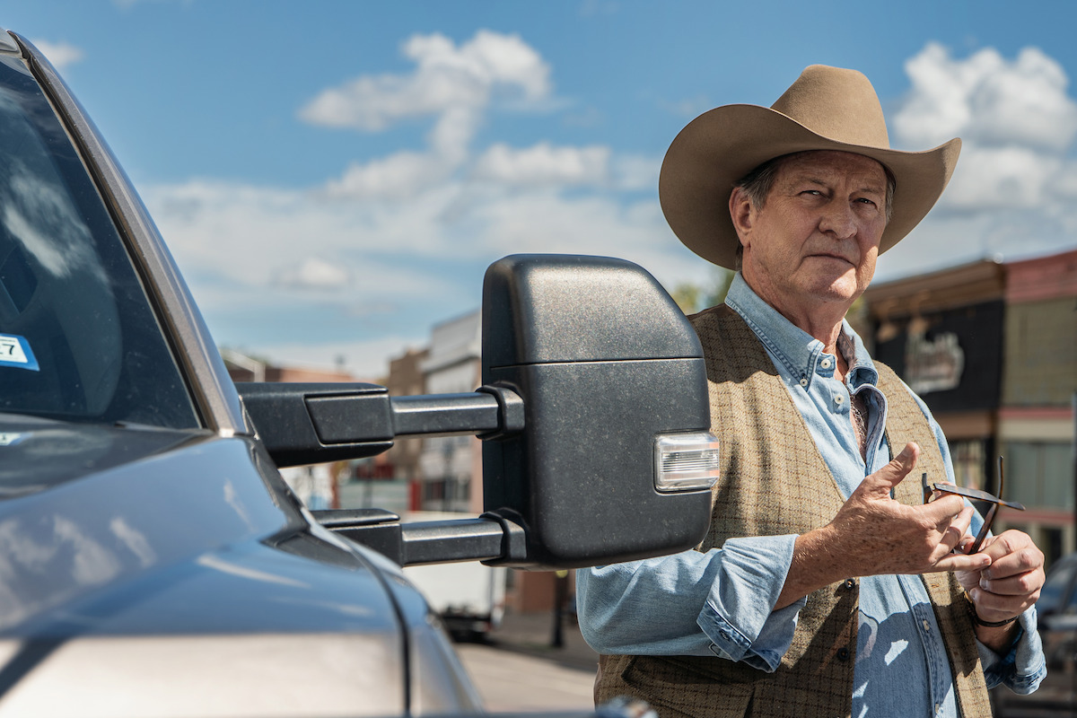 Older man in cowboy hat and vest stands by a pickup truck on a sunny day in a small town, buildings and blue sky with clouds in the background, holding sunglasses and looking thoughtful.
