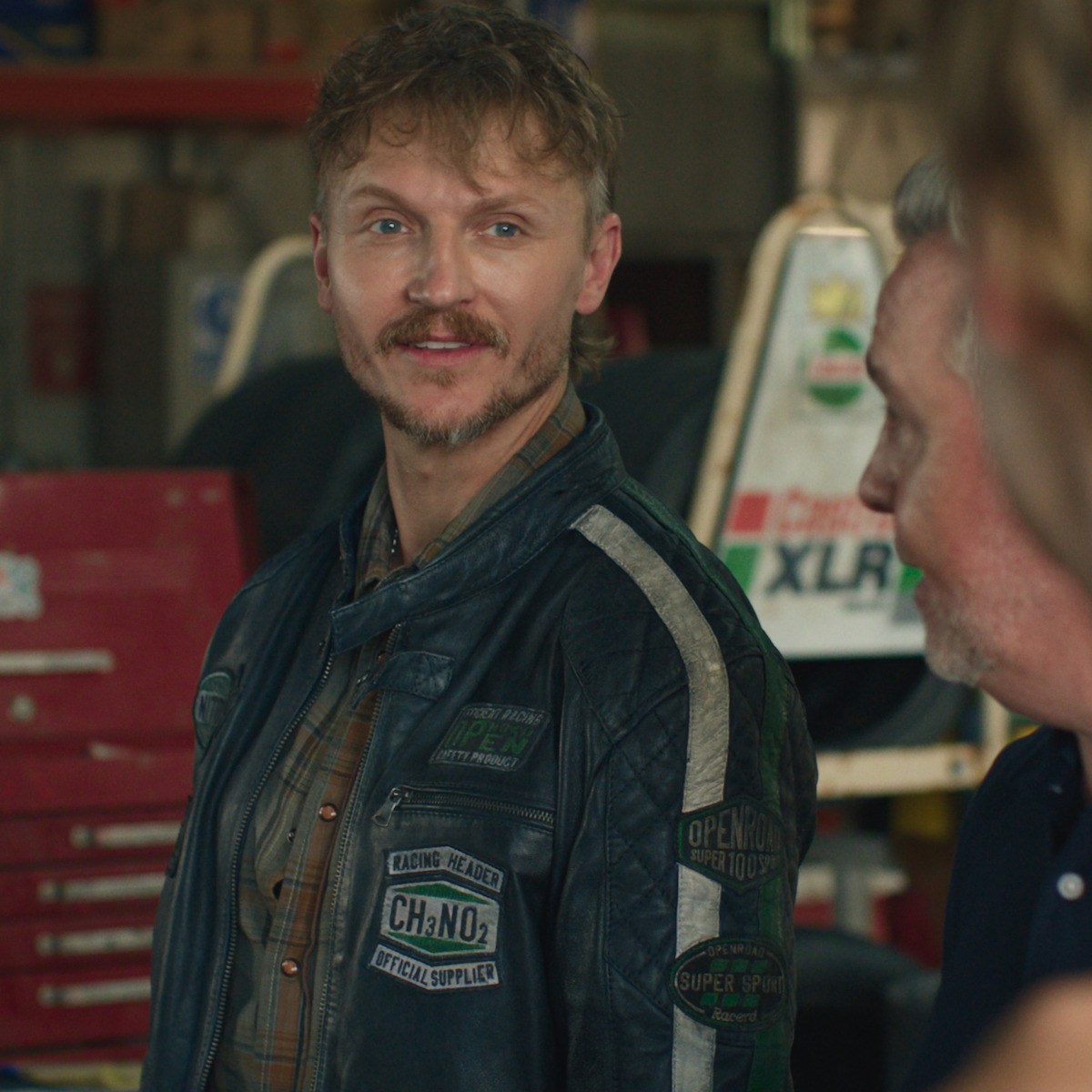 Man in a racing jacket standing in a garage talking with another man, surrounded by car parts, toolboxes, and automotive equipment in the background.