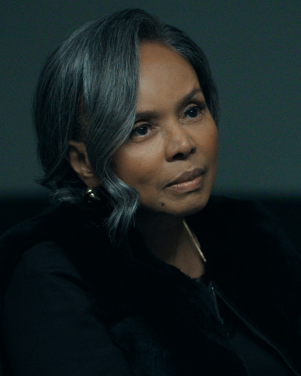 A woman with short gray hair in a dimly lit indoor setting, wearing a dark outfit and gold earrings, looking thoughtful and calm.