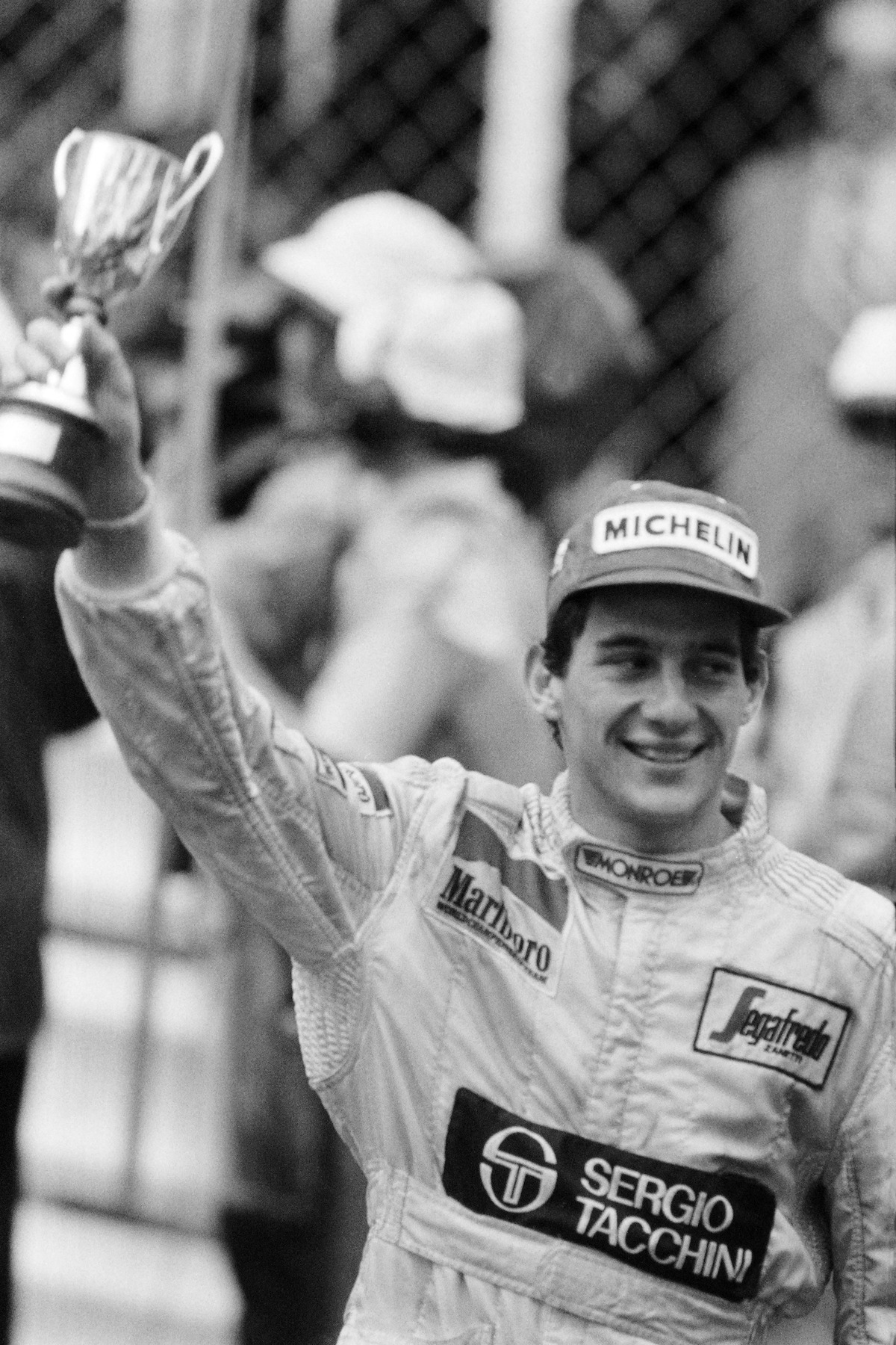 A black-and-white photo of Senna lifting his second place trophy at the Monaco Grand Prix in 1984. He has a big smile across his face.