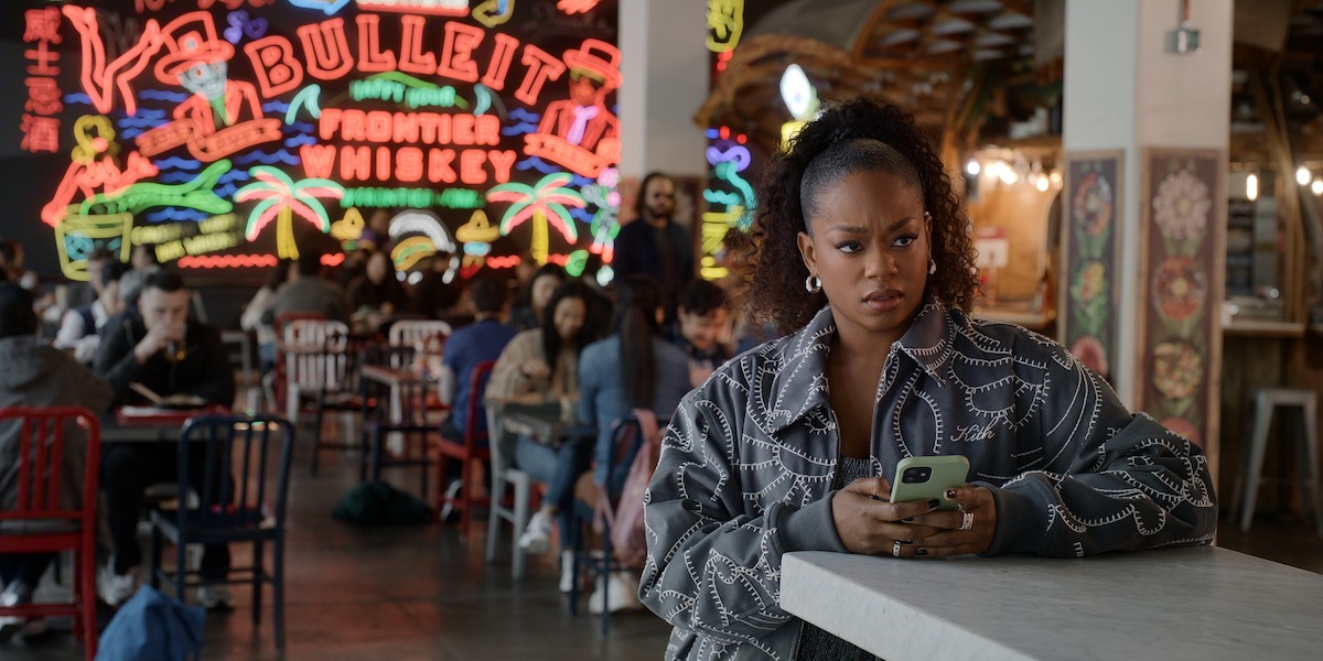 Woman in patterned jacket holding phone looks concerned at a counter in a busy cafe or food court with neon Bulleit Whiskey sign and people sitting at tables in the background.
