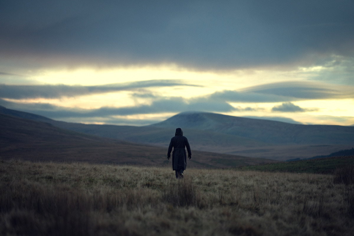 A solitary person in a dark coat walks across an open grassy field toward distant hills under a cloudy, moody sky at dusk. The landscape is vast and empty, evoking a sense of solitude and isolation.