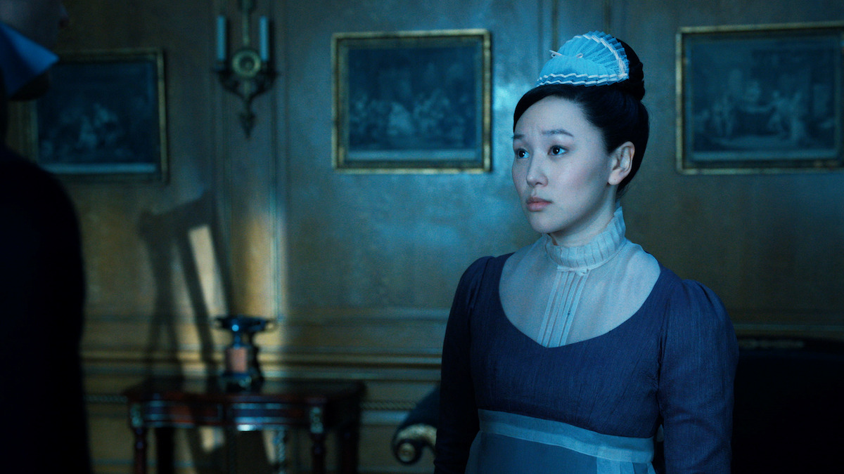 A woman in period costume and bonnet stands in a dimly lit, elegant room with wooden walls, framed paintings, and antique furniture, looking thoughtfully toward a person partially out of frame.