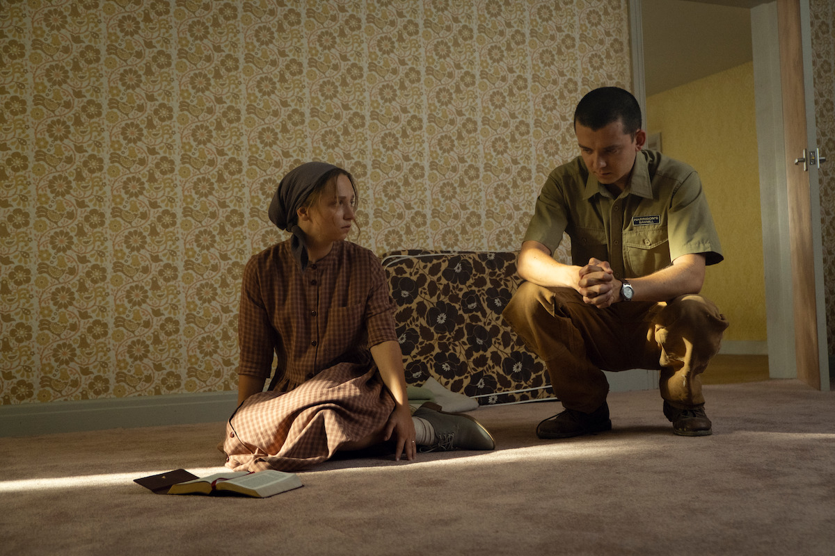 A woman in traditional clothing sits on a carpeted floor next to a book while a man in modern attire kneels beside her. They are in a sparsely furnished room with patterned wallpaper and soft lighting.