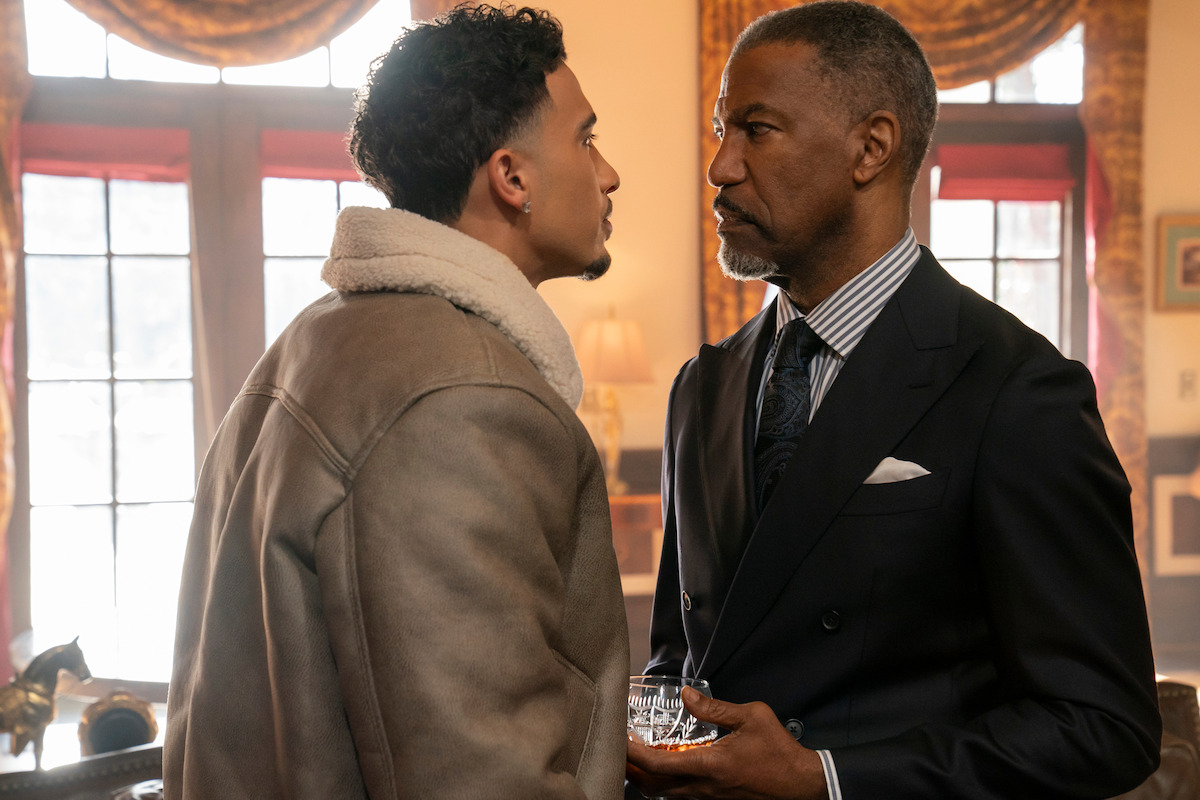 Two men stand face to face in an upscale, warmly lit room with large windows and elegant curtains, one in a suit holding a drink, the other in a shearling jacket, both appearing tense and engaged in a serious confrontation.