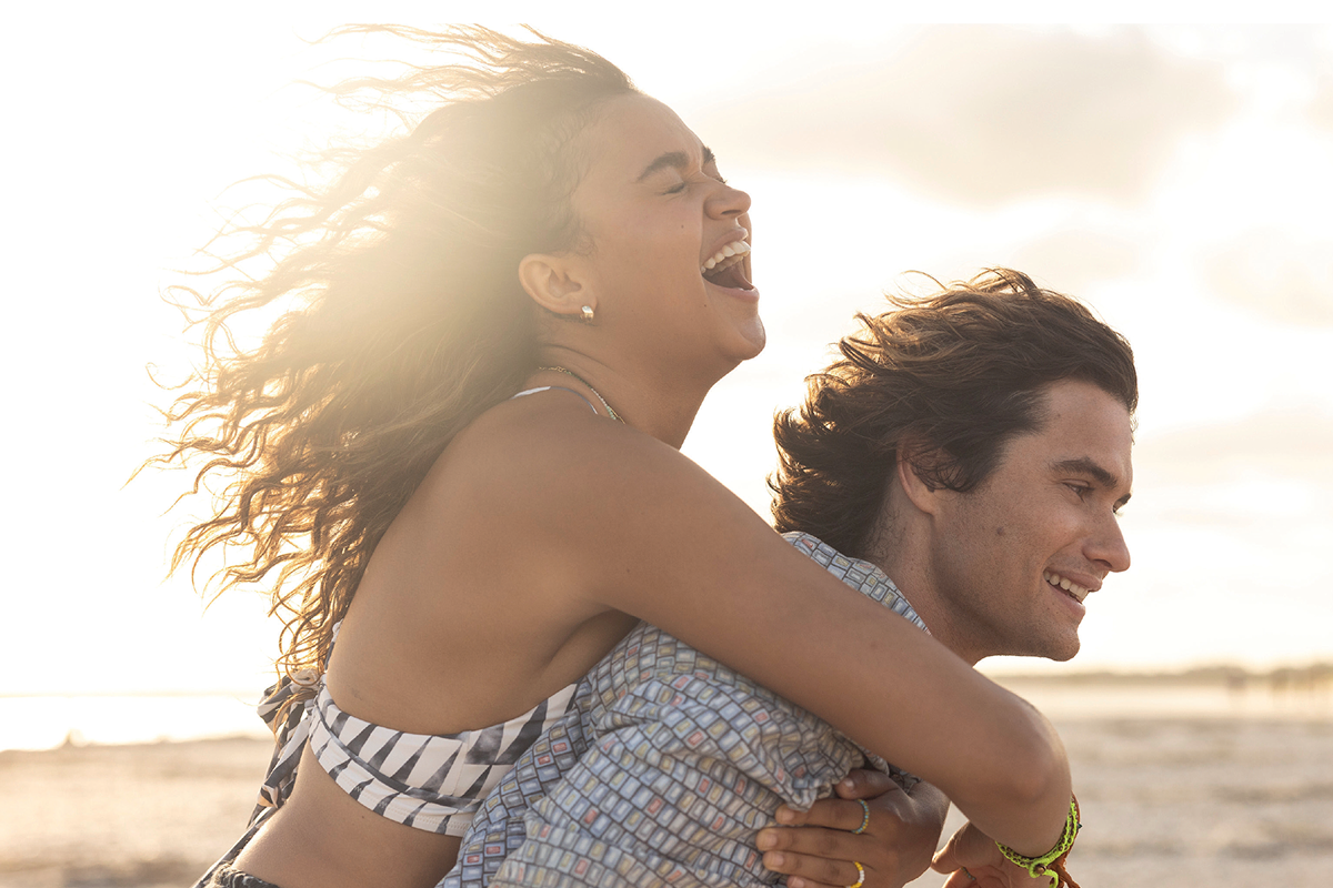 Two young people enjoying a piggyback ride on a sunny beach, both smiling and laughing with wind in their hair, relaxed and happy in a natural, outdoor setting with the ocean and sky in the background.