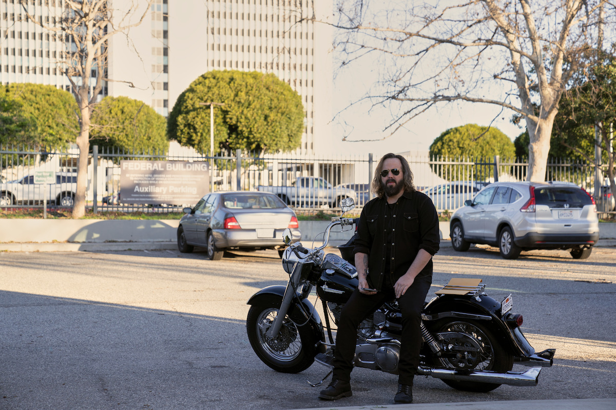 A man in dark clothing sits on a motorcycle in a parking lot with cars, trees, and a tall federal building in the background on a sunny day.