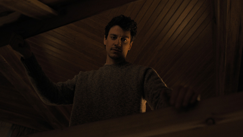 Man in a gray sweater standing indoors, looking down from a wooden loft or staircase area, with warmly lit wood-paneled ceiling in the background.