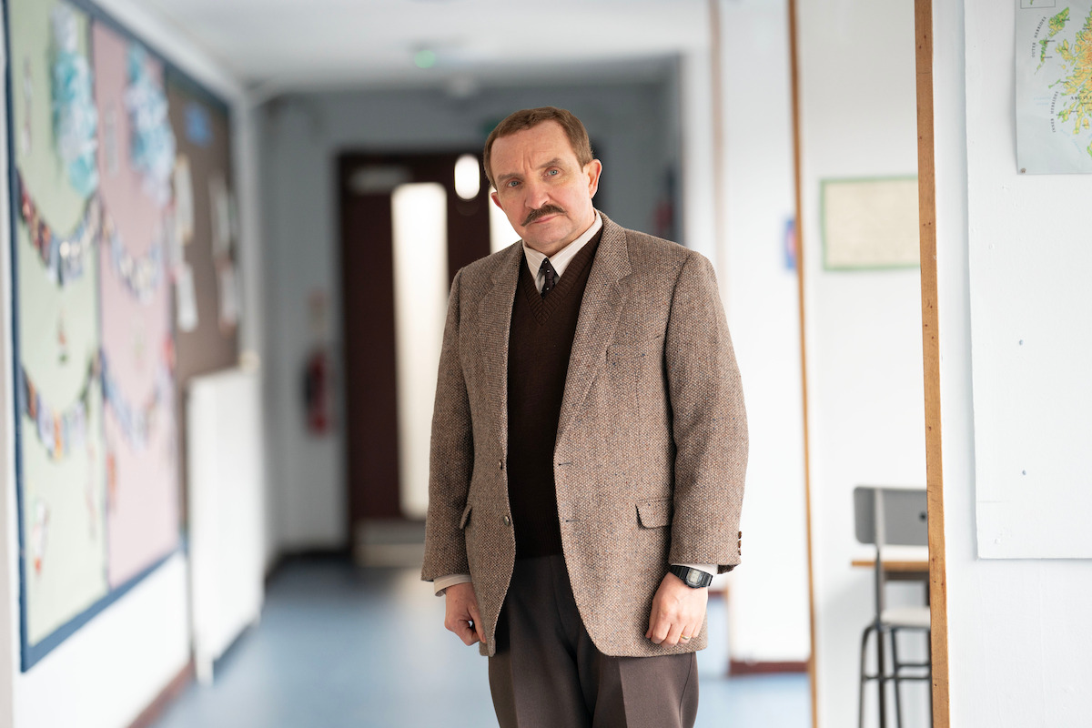 A man in a brown tweed jacket and tie stands in a well-lit school hallway, next to a bulletin board and map, with classroom doors and papers pinned on the walls in the background.