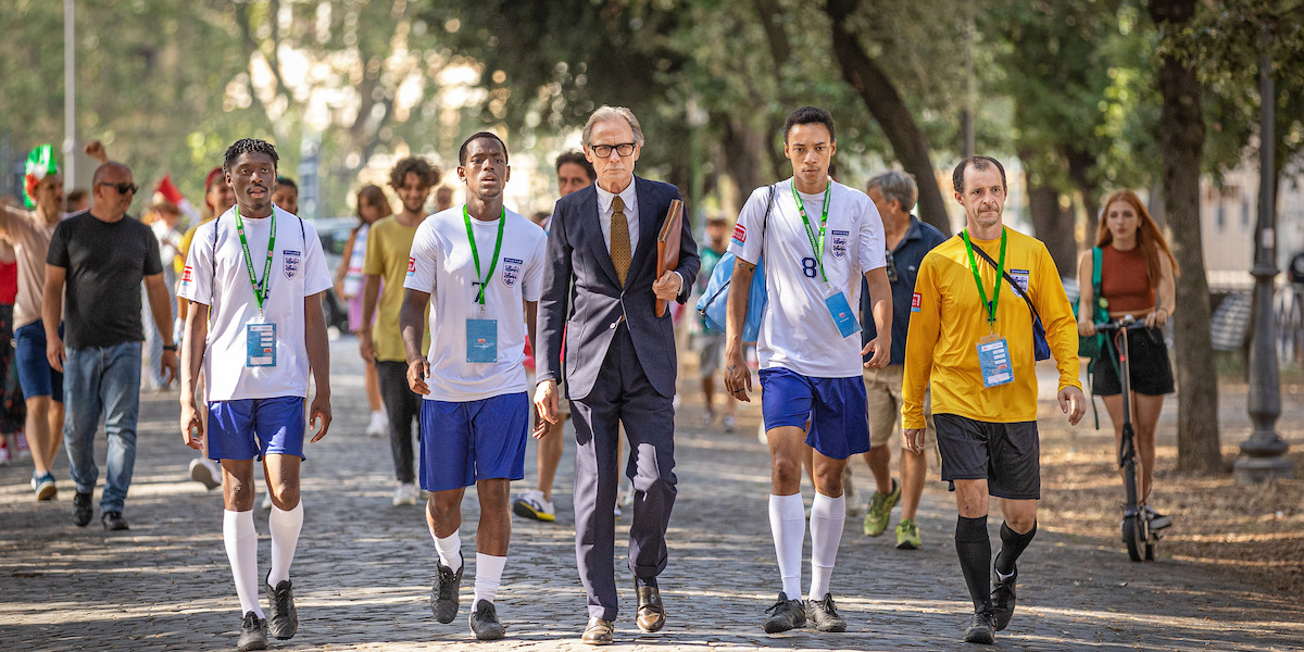 Man in suit walks alongside soccer players down a pedastrian path outside.