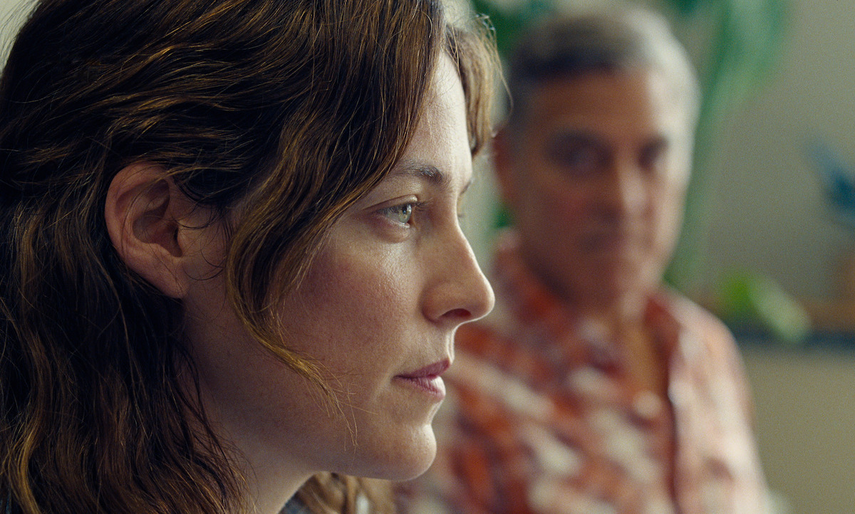 A woman in sharp focus looks thoughtful indoors, while a man sits in the blurred background. The lighting is natural and soft, creating a contemplative, quiet mood.