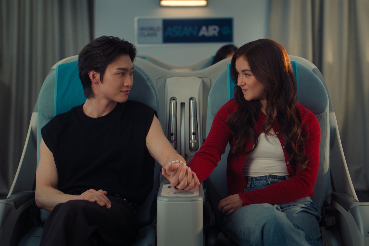 A young man and woman sitting side by side on an airplane, holding hands and smiling at each other; airplane seats and an "Asian Air" sign visible in the background.