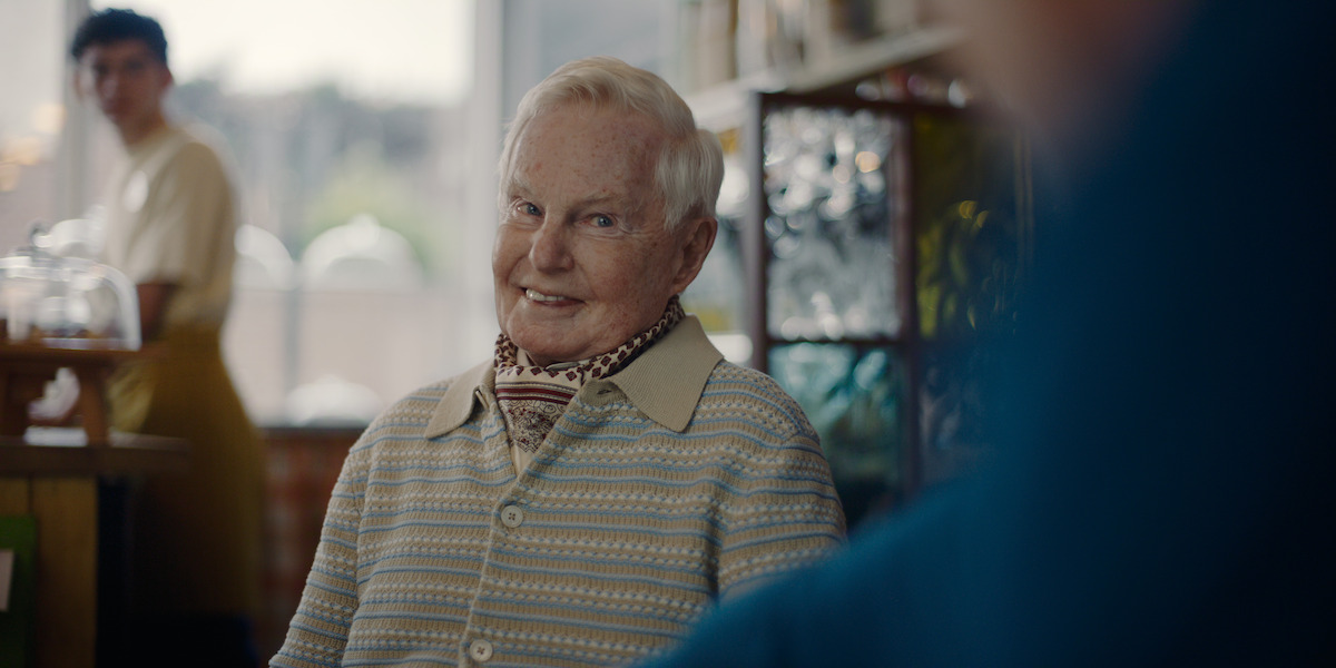 Elderly person smiling while seated in a cozy café, wearing a striped sweater and scarf. Soft light fills the room, with a person in the background near a counter and shelves, creating a warm, inviting atmosphere.
