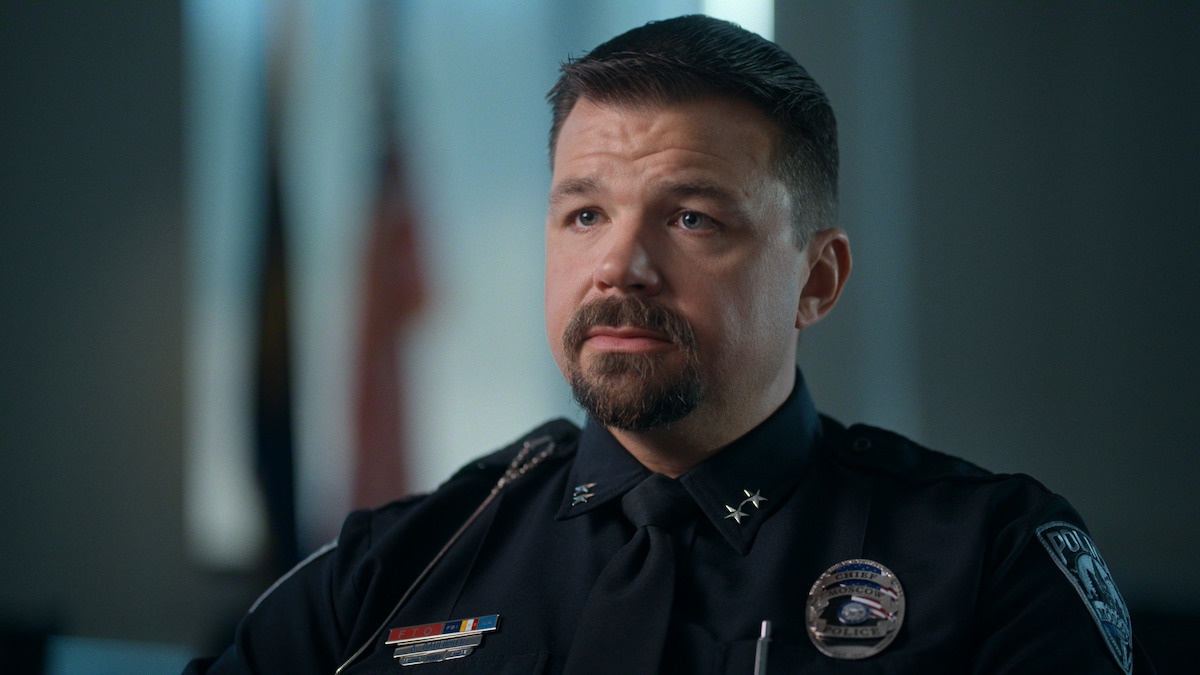 Police officer in uniform sitting indoors, with blurred flags and a softly-lit office background, looking serious and attentive.