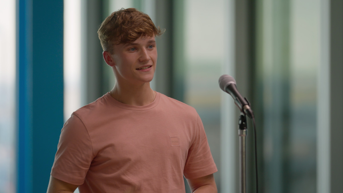 Young man in a pink shirt stands by a microphone indoors, modern glass building background, bright natural light, casual and confident mood.