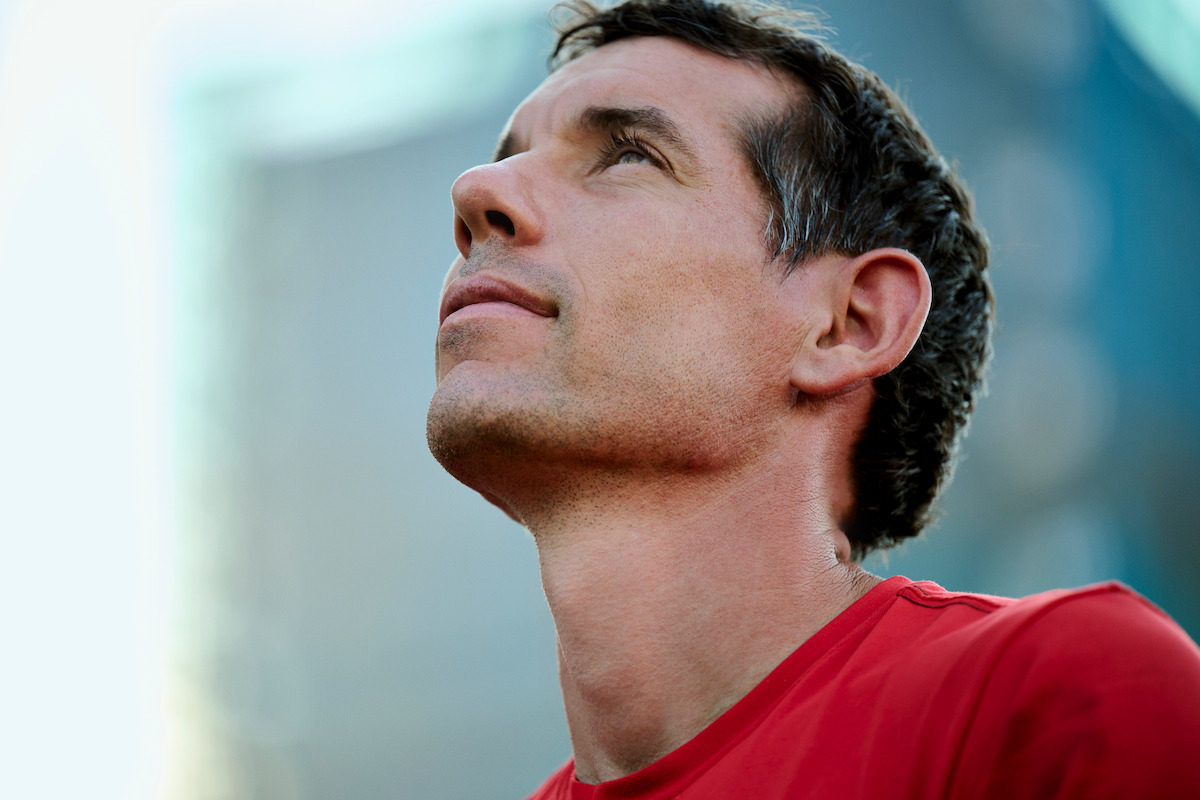 Alex Honnold in a red shirt looks upward with a thoughtful expression, outdoors with blurred urban background and sunlight.