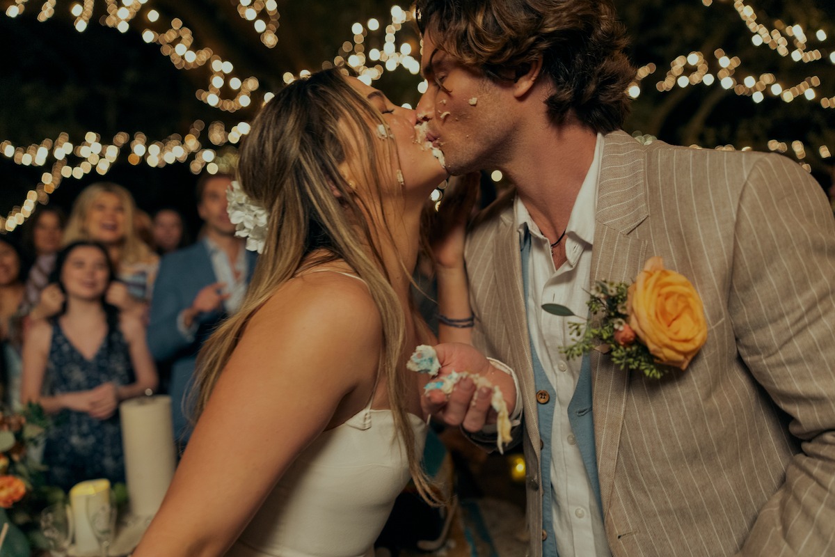Bride and groom kiss with cake on their faces at an outdoor wedding reception, surrounded by guests and festive string lights in the background.