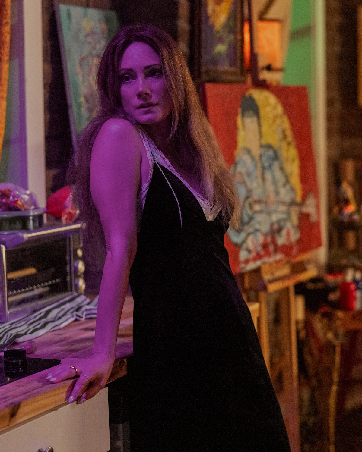 Natalie Brown como Ellie, de vestido preto, está encostada em uma bancada de cozinha, cercada por pinturas coloridas e materiais de arte em um estúdio artístico com iluminação aconchegante em tons de roxo.
