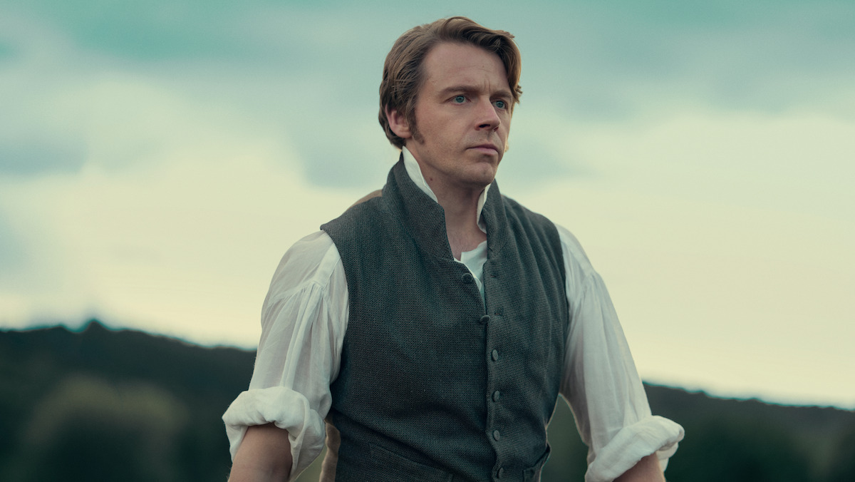 A man in historical clothing stands outdoors with a serious expression, wearing a vest over a white shirt, with hills and trees in the blurred background under a cloudy sky.