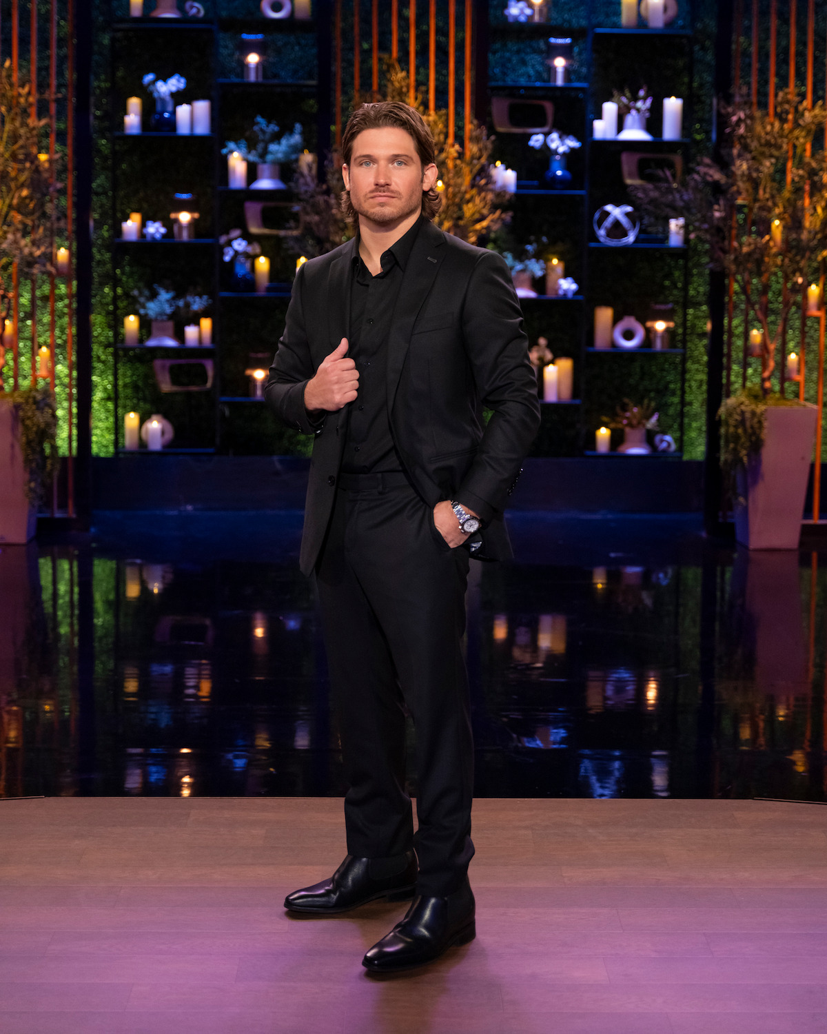 A man in a black suit stands on a stage with a wood floor and decorated background shelves featuring candles, plants, and vases with warm lighting, creating an elegant and formal atmosphere.