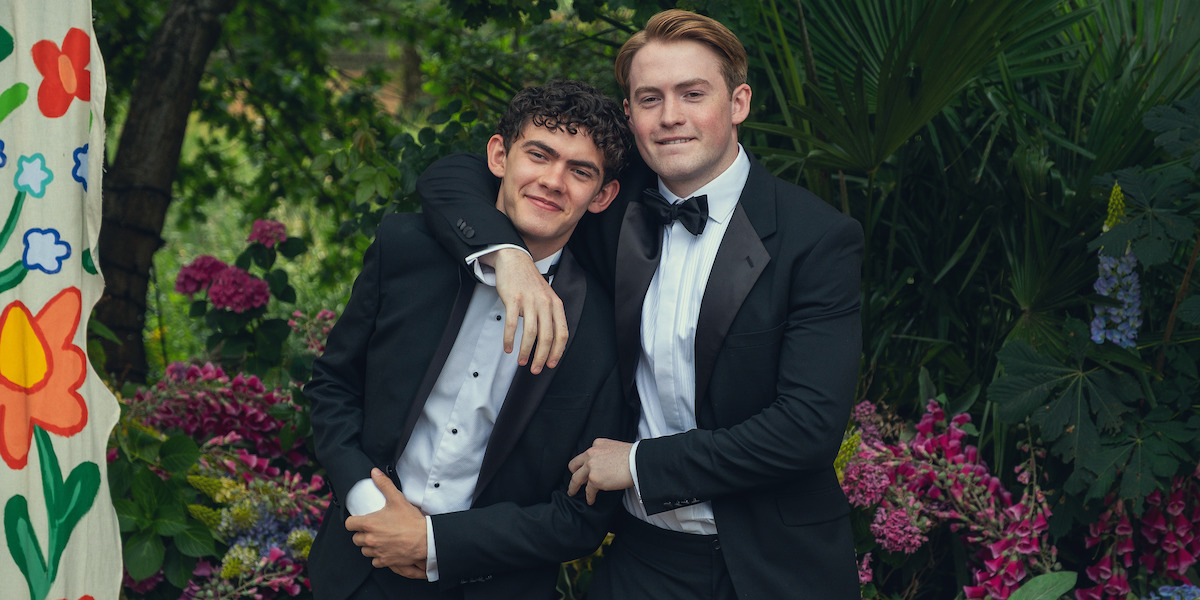 Two young men in black tuxedos pose together outdoors, standing close with one arm around the other, smiling in a lush garden surrounded by vibrant flowers and greenery, with a colorful floral backdrop to the left.
