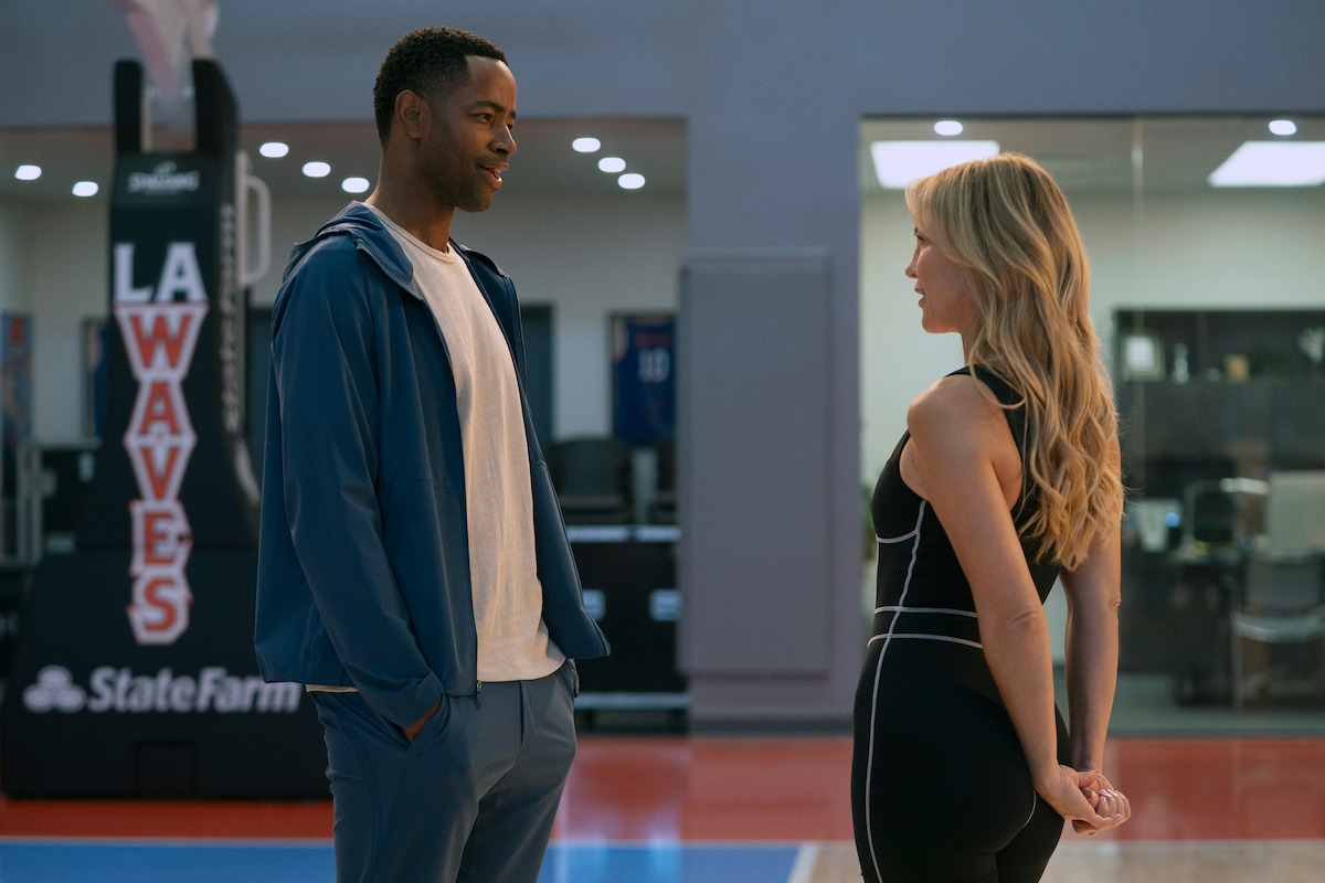 A man and woman stand and talk on an indoor basketball court near a hoop with "LA Waves" signage, in a modern gym setting with office space visible in the background.