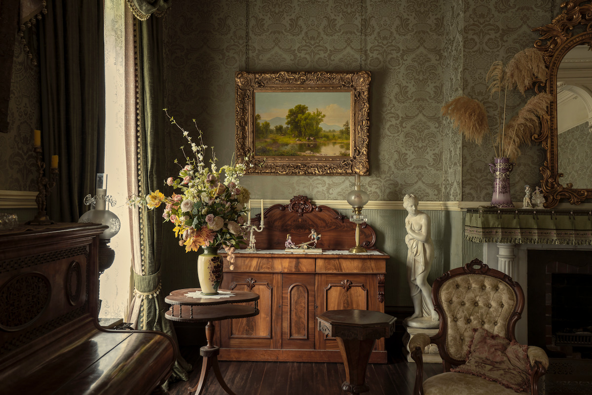 Elegant vintage living room with floral arrangement, ornate furniture, patterned wallpaper, framed landscape painting, fireplace with decor, statues, and soft, natural lighting from tall draped windows.