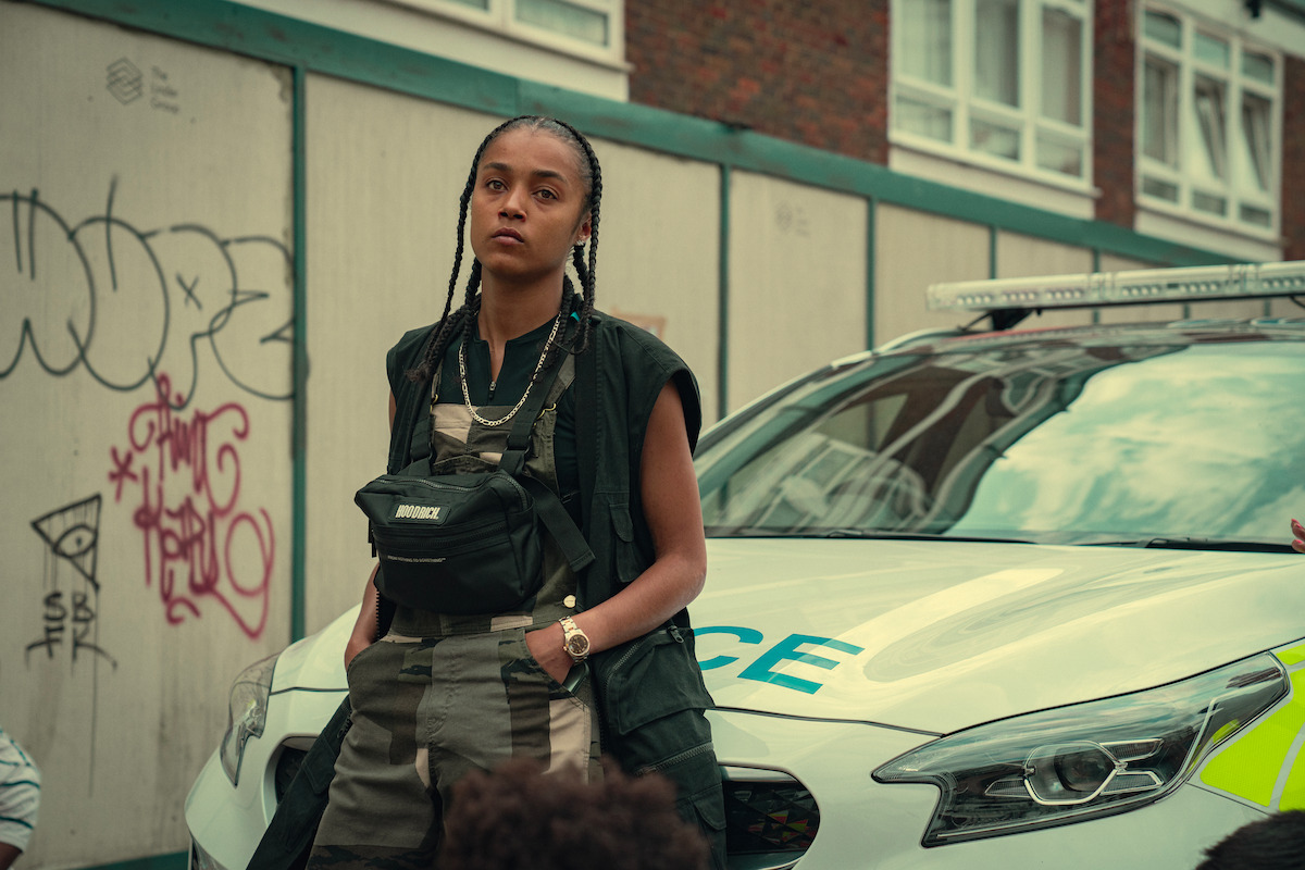 A woman with braids leans against a police car on a city street, wearing a black vest and utility bag, with graffiti and apartment buildings visible in the background. Urban setting with a serious mood.