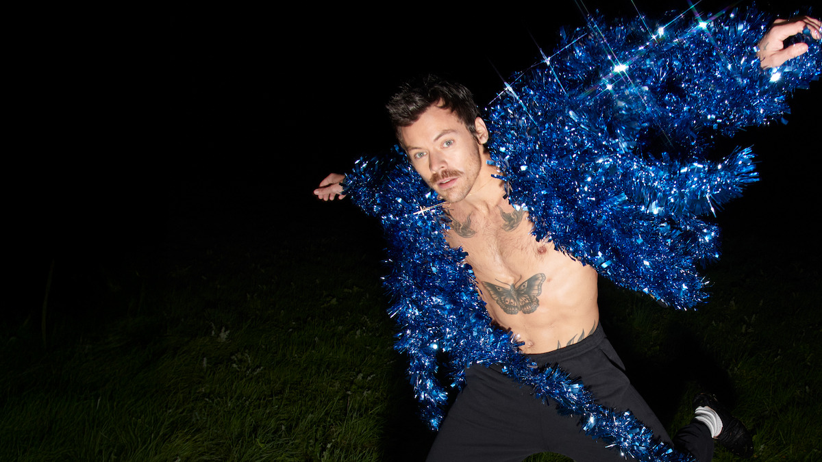 Man with mustache posing outdoors at night, wearing a sparkling blue tinsel jacket open to show his chest and tattoos, standing on grass with a dark background, in a dramatic and playful stance.