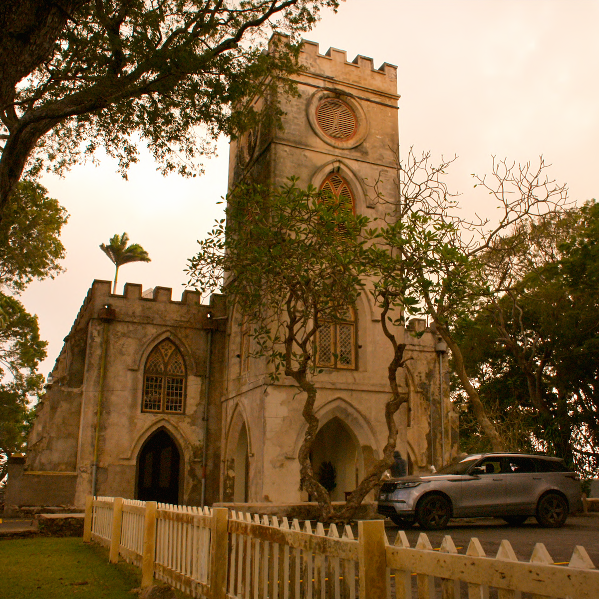 Igreja histórica de pedra com janelas em arco e torres, cercada por árvores e uma cerca de madeira. Há um carro estacionado na frente dela. O céu está nublado e a iluminação é suave, do início da noite.