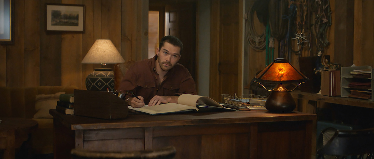 Man in a brown shirt sits at a wooden desk in a rustic, warmly lit office, writing in a large book. Lamps, ropes, books, and wood-paneled walls create a cozy, ranch-like environment.