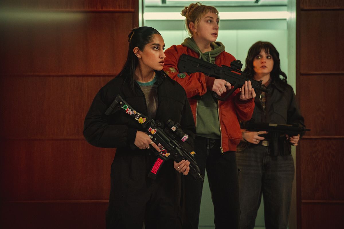 Três mulheres de roupas informais ficam em estado de alerta e seguram armas de brinquedo no corredor de um prédio moderno, com um elevador e paredes de madeira ao fundo. Elas olham atentas para os lados.