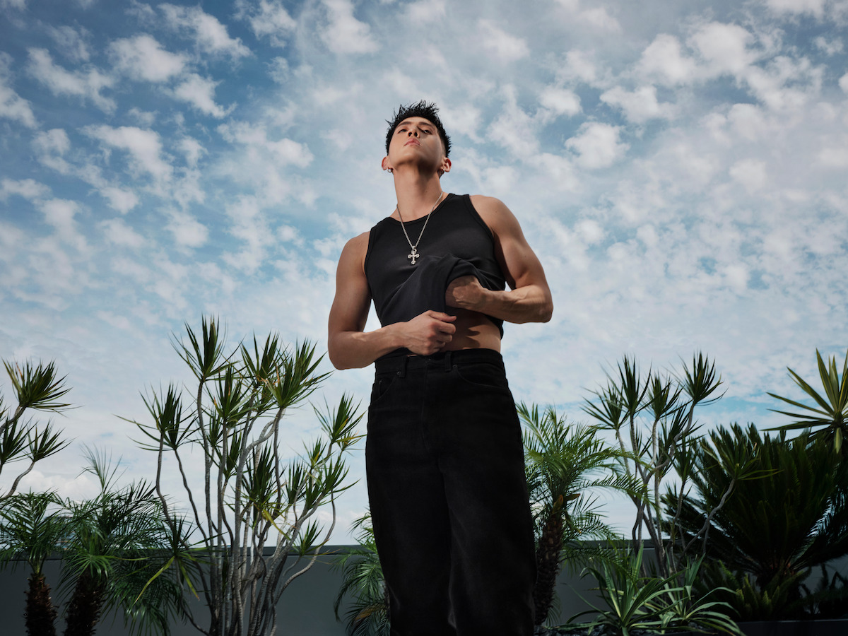 Man in black sleeveless shirt and dark pants standing outdoors among palm plants, looking up at a cloudy sky, with modern landscaping visible in the background.